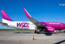 როგორ ვიმოგზაუროთ ქუთაისიდან – Wizz Air A side profile view of a Wizz Air Airbus A320 aircraft with its white, purple, and magenta livery, parked on the airport tarmac under a clear blue sky. lui.ge