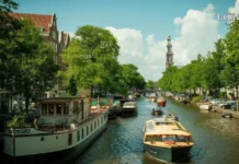 ამსტერდამი – ევროპული თავისუფლებისა და არხების მაგია: სრული გზამკვლევი Tourist boats and houseboats on an Amsterdam canal lined with green trees and historic buildings under a blue sky.